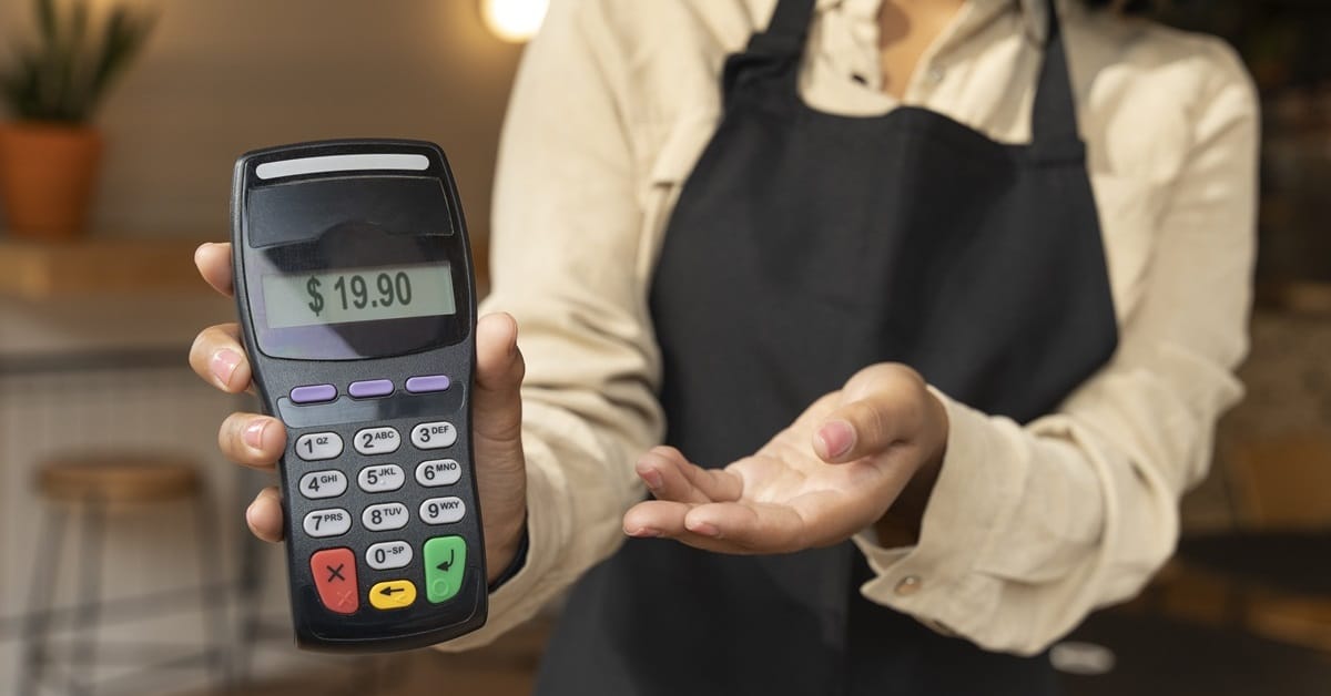 Pessoa segurando uma máquina de cartão exibindo o valor $19,90, com foco no equipamento e fundo desfocado de ambiente comercial.