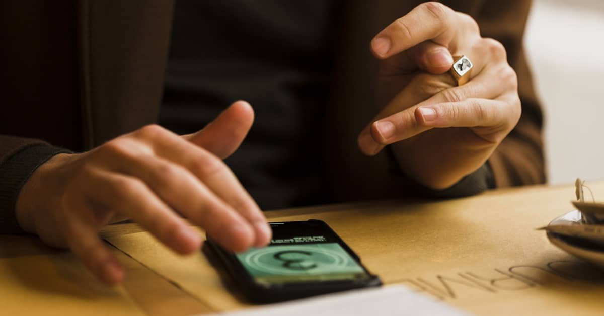 Pessoa utilizando smartphone em uma mesa de madeira para realizar pagamento digital, com tela mostrando símbolo de moeda e interface tecnológica