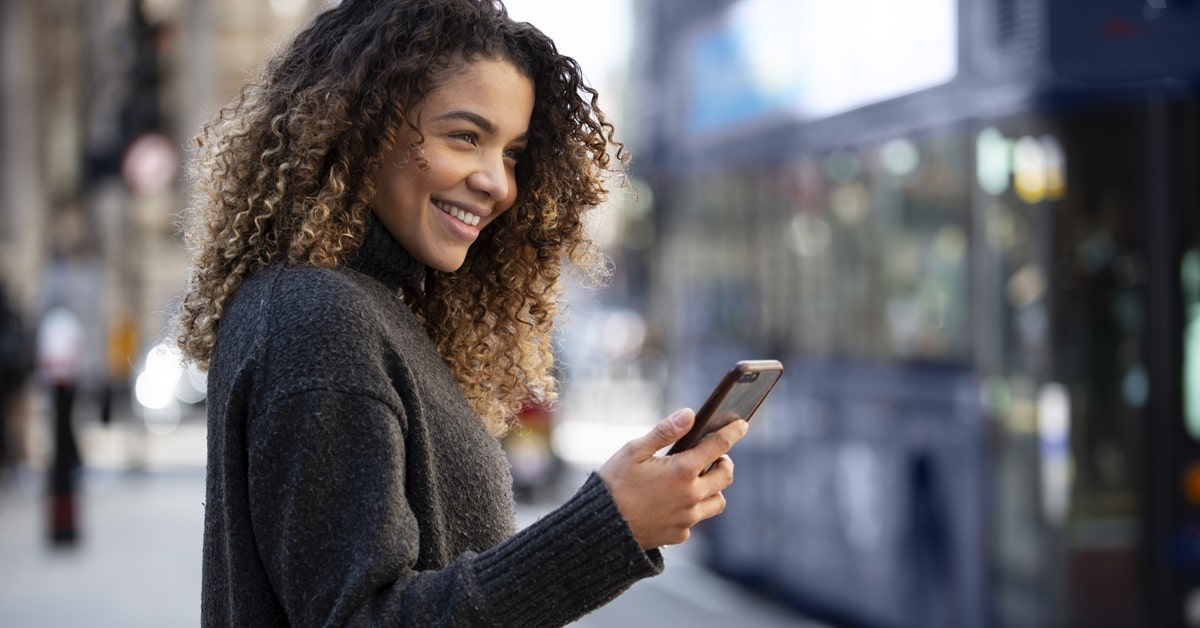 Mulher sorridente utilizando smartphone em um ambiente urbano, representando praticidade e tecnologia em pagamentos digitais.