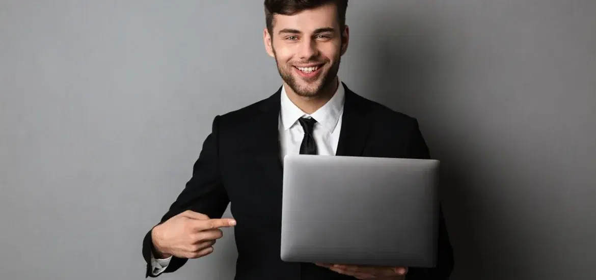 Jovem profissional de terno segurando um laptop, sorrindo e apontando para a tela em ambiente corporativo com fundo cinza.