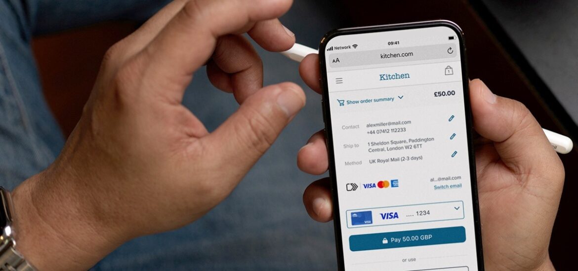 Mão segurando smartphone exibindo a tela de pagamento online com a opção dos avanços no Click to Pay e informações de contato e envio.