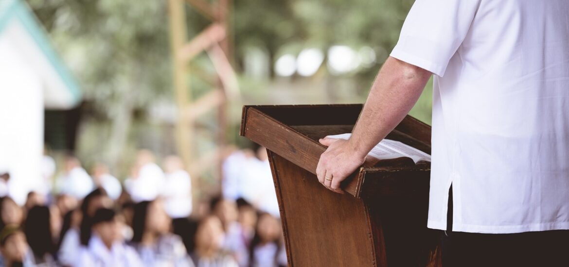 Pregador de camisa branca discursando em um púlpito de madeira em um ambiente ao ar livre com público ao fundo desfocado.