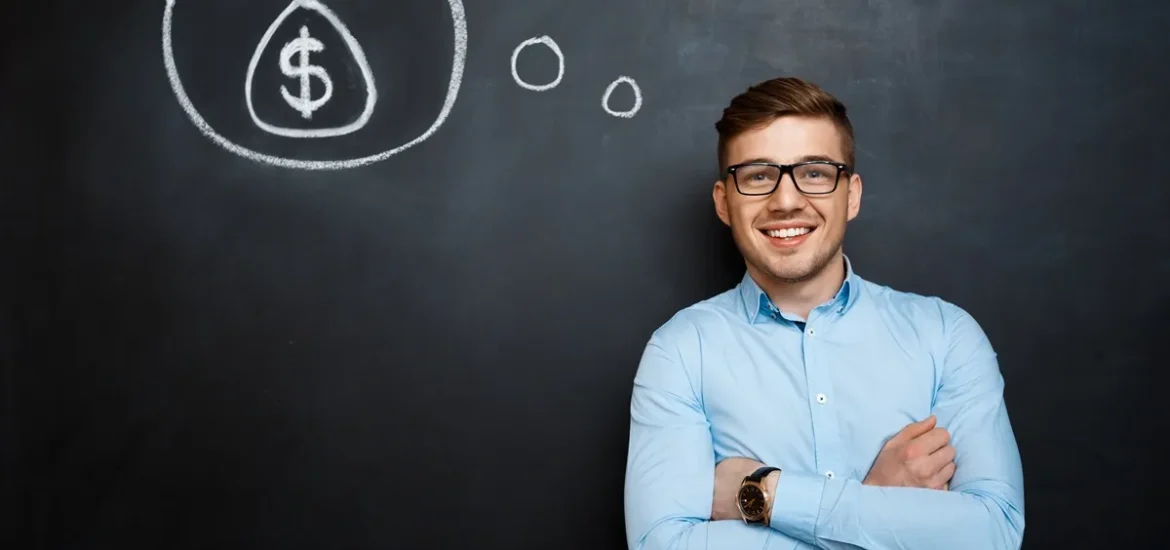 Homem sorridente com camisa azul, posicionado ao lado de um quadro-negro com um desenho de um saco de dinheiro dentro de um balão de pensamento.