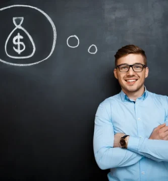 Homem sorridente com camisa azul, posicionado ao lado de um quadro-negro com um desenho de um saco de dinheiro dentro de um balão de pensamento.