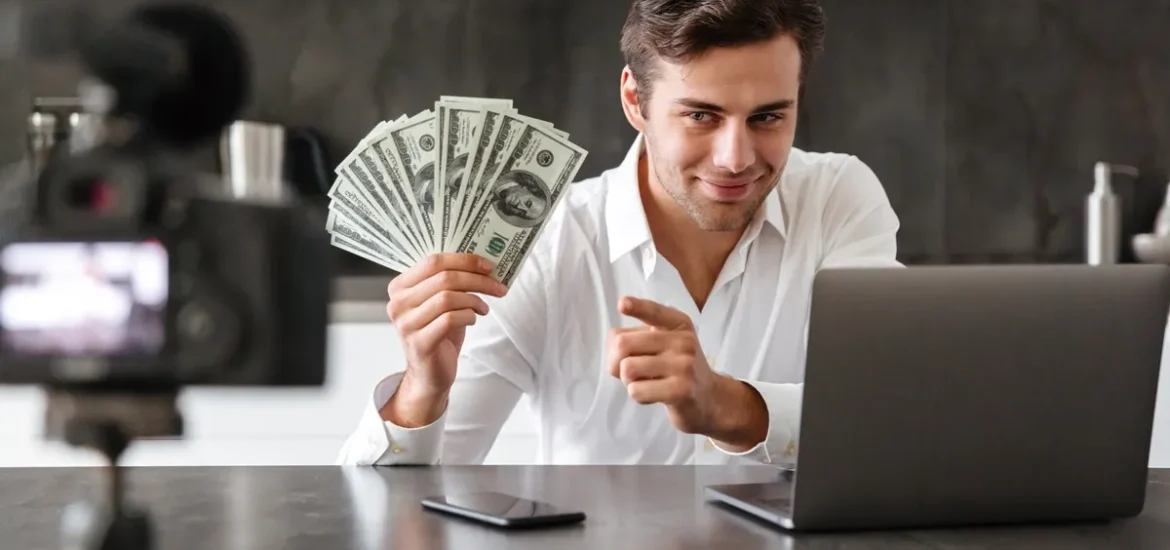 Homem sorridente segurando notas de dólares enquanto grava um vídeo com câmera e laptop, simbolizando geração de renda online e marketing digital.