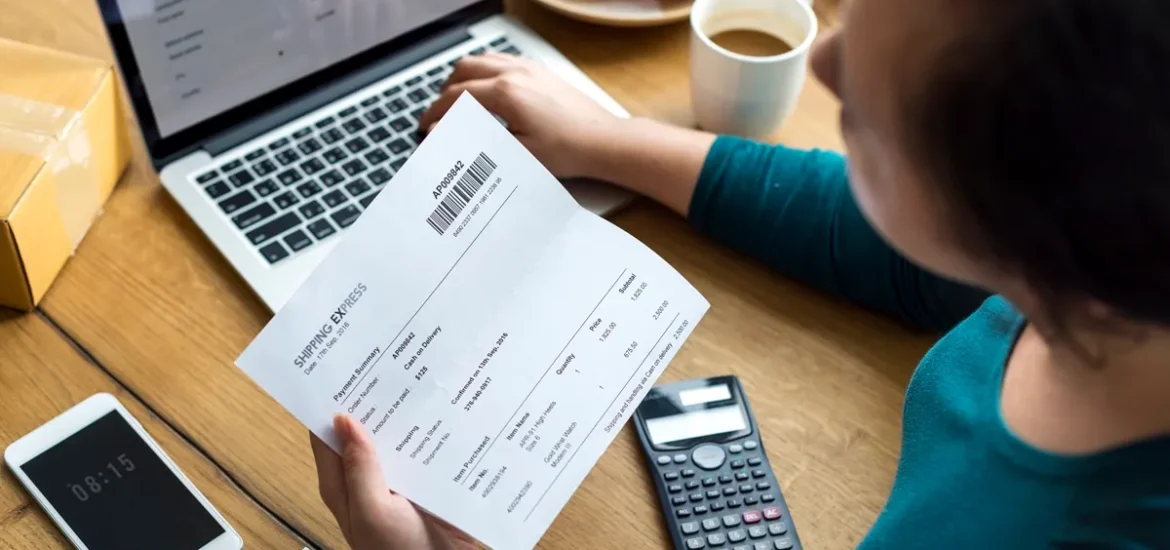 Pessoa analisando uma fatura em papel enquanto utiliza um laptop para verificação de pagamento online.