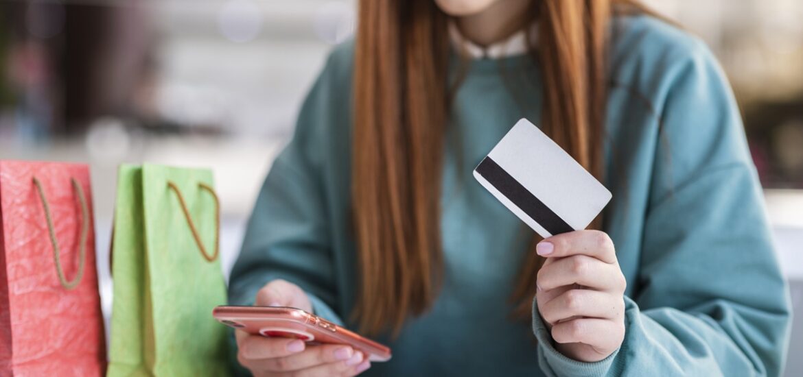 Mulher segurando um cartão de crédito enquanto utiliza um smartphone, com sacolas de compras ao fundo, simbolizando segurança e praticidade em compras online