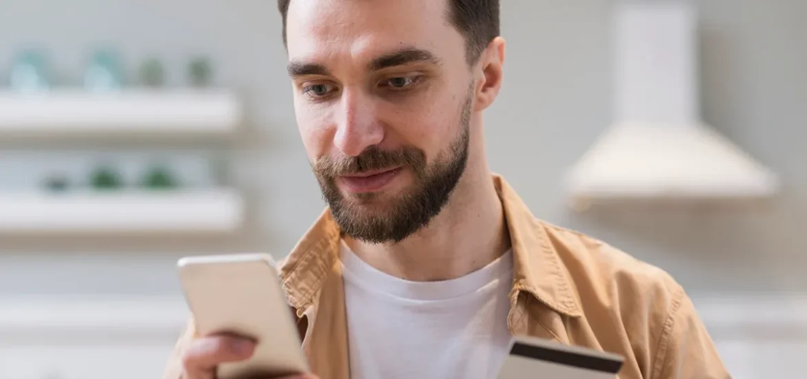 Homem segurando um smartphone e um cartão de crédito, realizando uma transação em um ambiente interno.