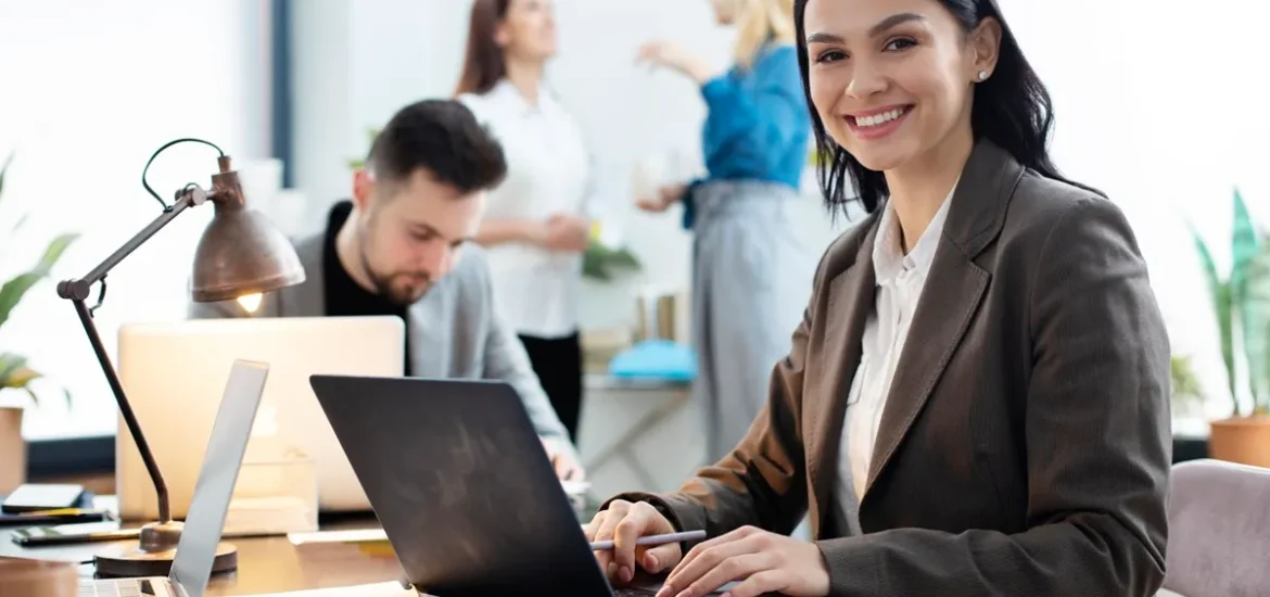 Mulher sorridente trabalhando em um escritório moderno, com colegas ao fundo em um ambiente colaborativo e produtivo.