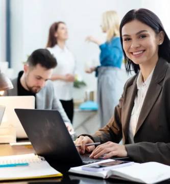 Mulher sorridente trabalhando em um escritório moderno, com colegas ao fundo em um ambiente colaborativo e produtivo.