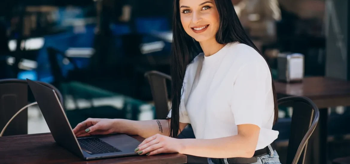 Mulher sorridente trabalhando em um laptop em uma mesa ao ar livre, com um ambiente casual e descontraído ao fundo.