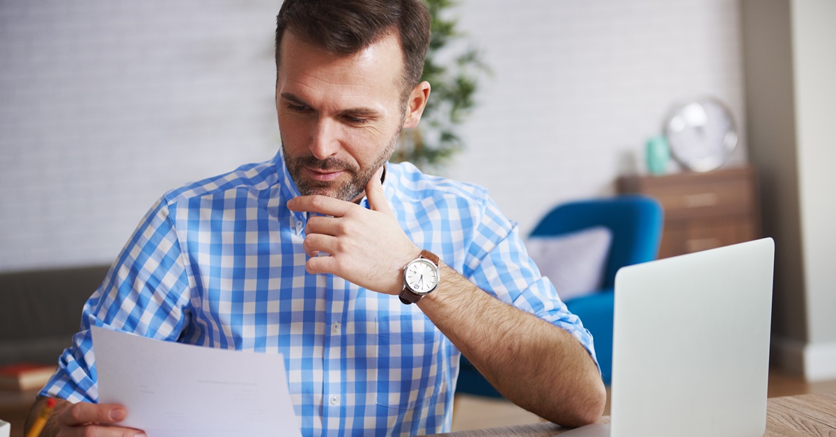 Homem de meia-idade, vestindo camisa xadrez azul e branca, examina um documento em casa ou no escritório, com um laptop aberto à sua frente e uma decoração moderna ao fundo