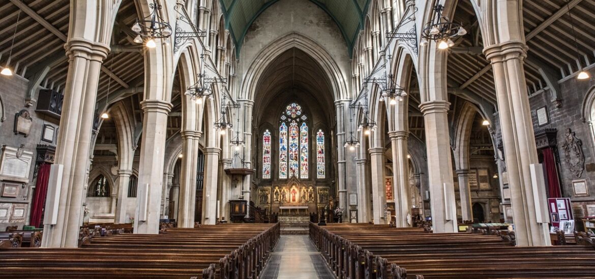 Interior de uma igreja com arquitetura gótica, bancos de madeira alinhados e vitrais coloridos ao fundo.