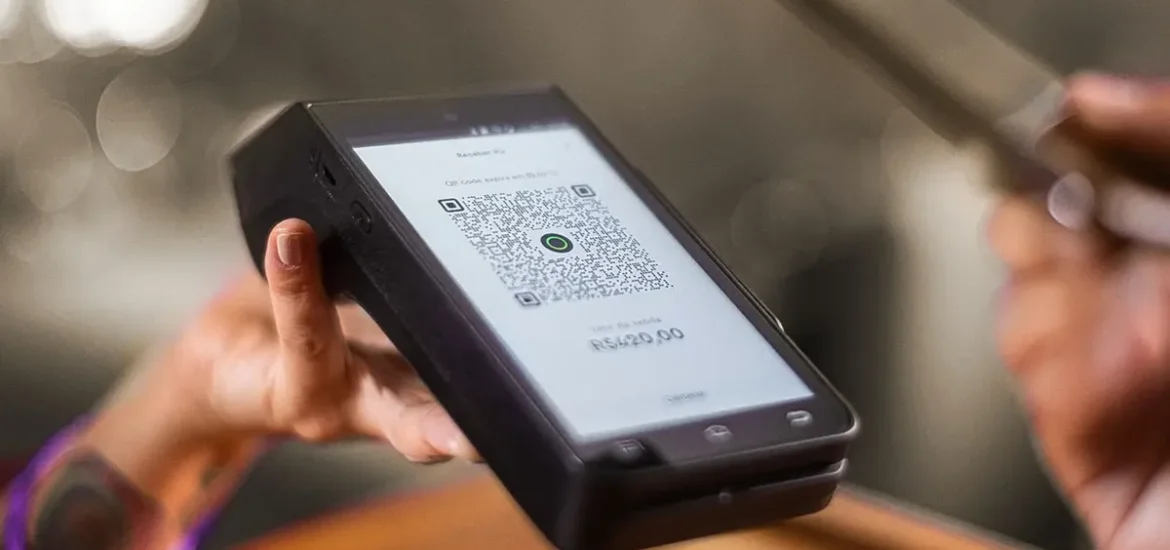 Pessoa segurando maquininha de cartão exibindo QR Code para pagamento via Pix no valor de R$ 420,00.