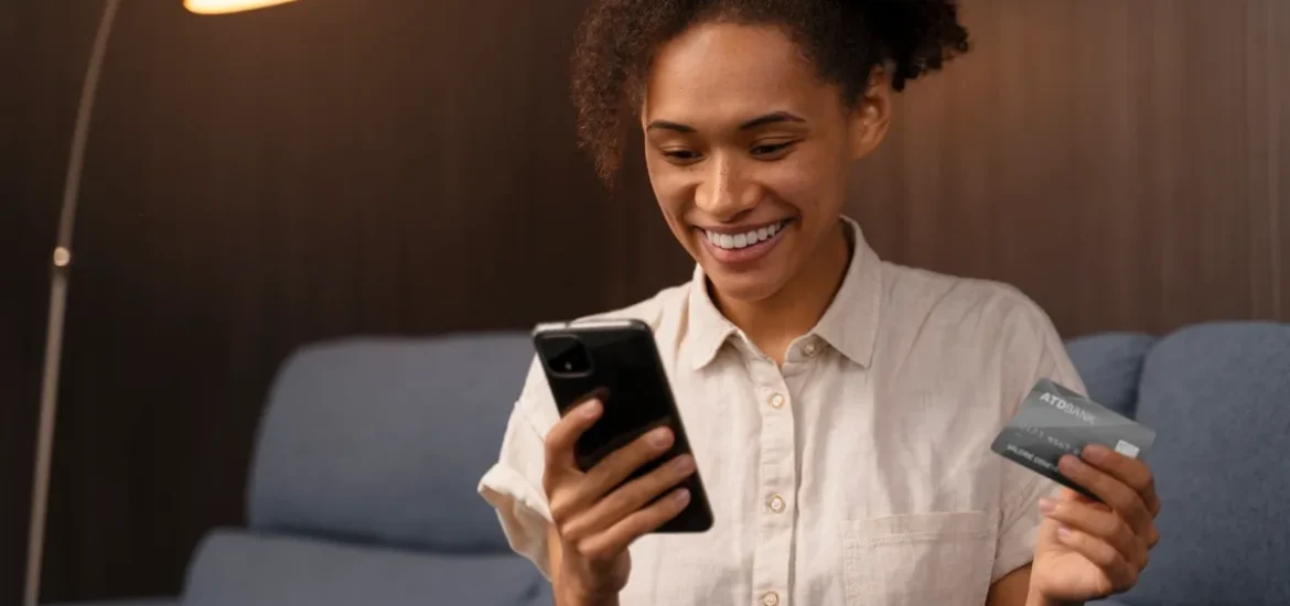 Mulher sorridente segurando um smartphone e um cartão de crédito, realizando uma transação em ambiente aconchegante.