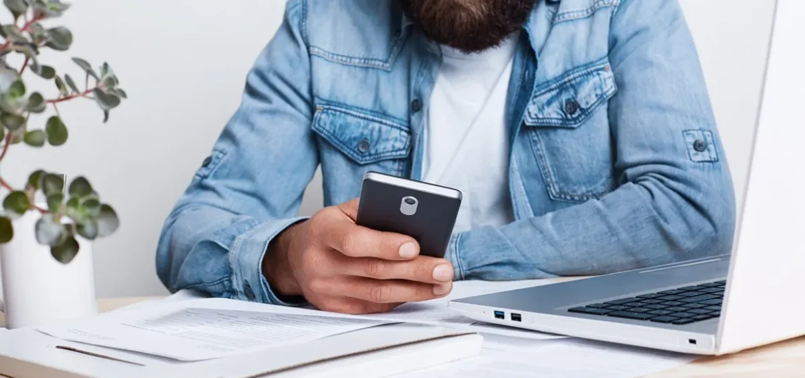 Homem com camisa jeans, utilizando um smartphone em uma mesa de trabalho com documentos e notebook.