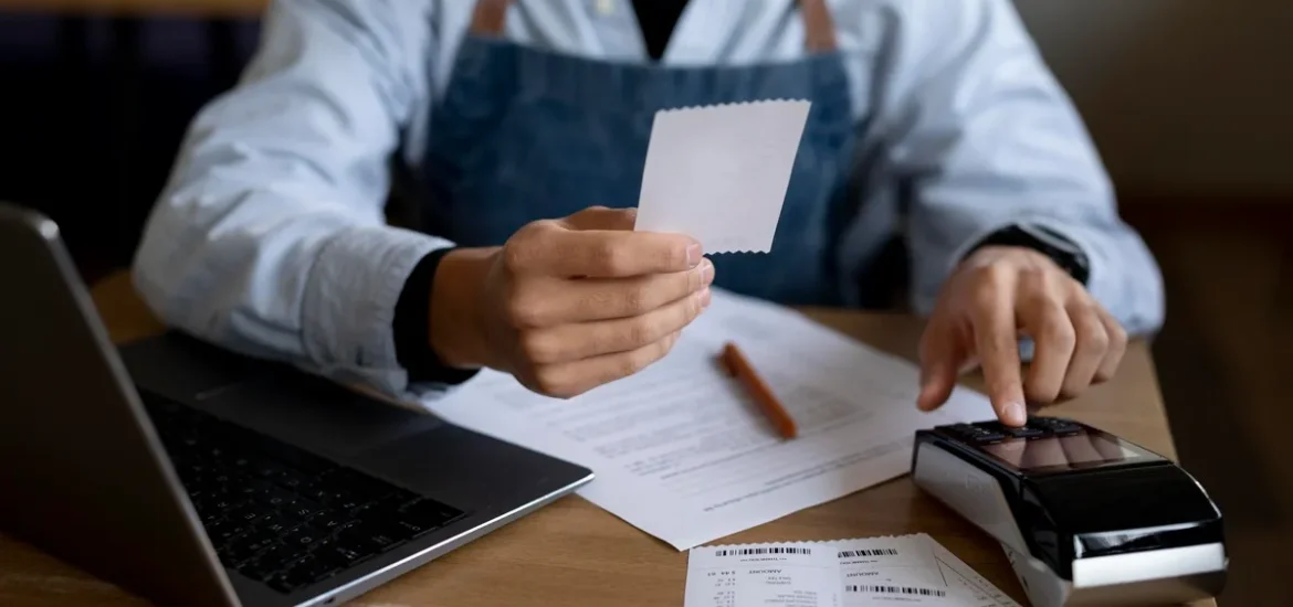 Pessoa com avental azul analisando um recibo em frente a uma máquina de cartão e laptop em uma mesa de trabalho.