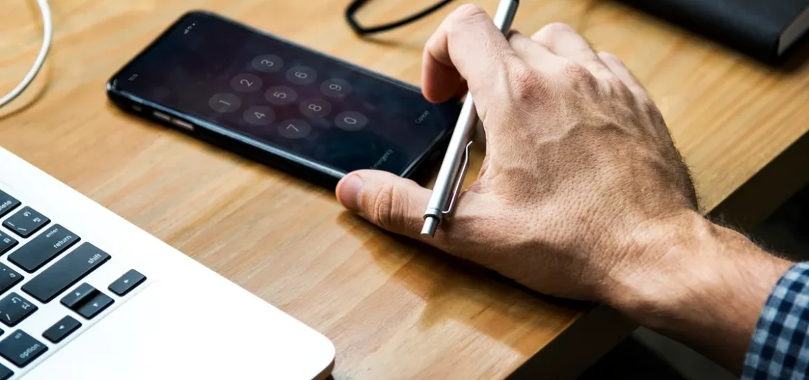 Mão segurando uma caneta sobre um smartphone desbloqueado, em uma mesa de trabalho com notebook ao lado.