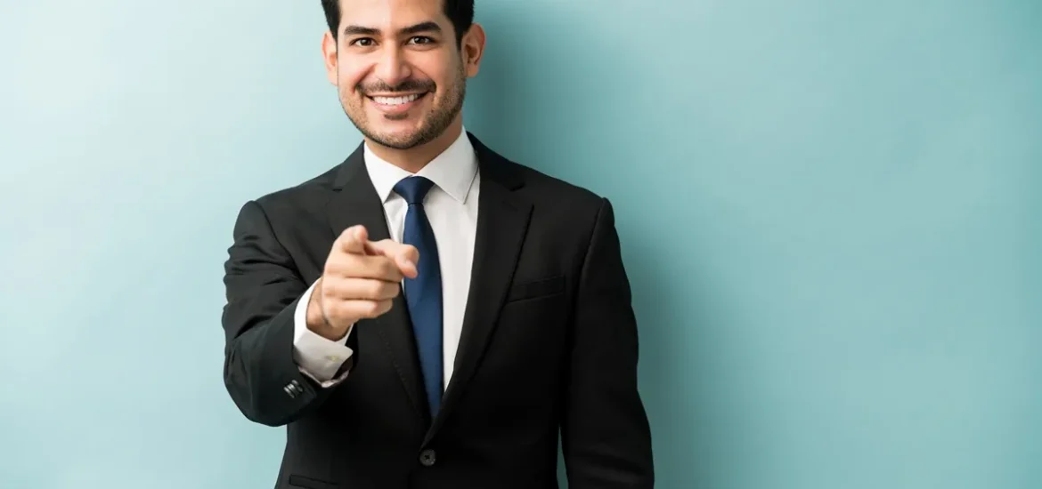 Homem de terno preto e gravata azul, sorrindo confiante enquanto aponta para a câmera, com fundo azul claro