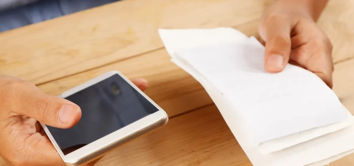 Mãos segurando um smartphone e uma pilha de recibos em uma mesa de madeira.