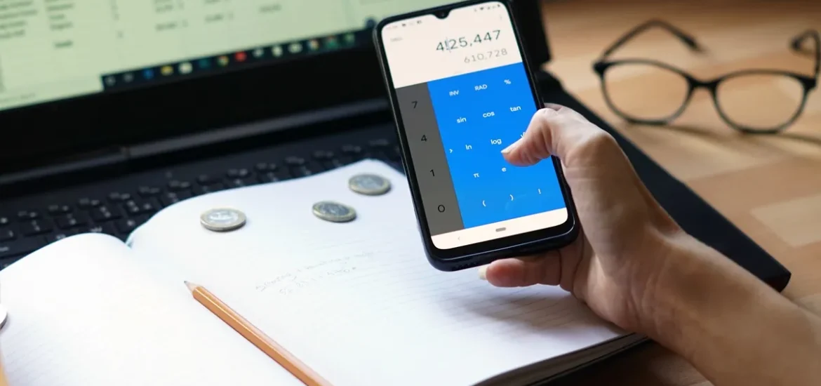 Mão segurando um smartphone com calculadora aberta, ao lado de um notebook, caderno e moedas sobre a mesa.