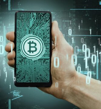 Mão segurando um smartphone com símbolo de Bitcoin na tela, representando pagamentos digitais e integração entre criptomoedas e sistemas financeiros tradicionais. No fundo, códigos binários e uma cidade digital simulam o conceito de blockchain e transações digitais seguras.