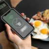 Pessoa realizando pagamento via NFC em smartphone enquanto um prato de café da manhã com ovos, bacon e salada está em segundo plano.