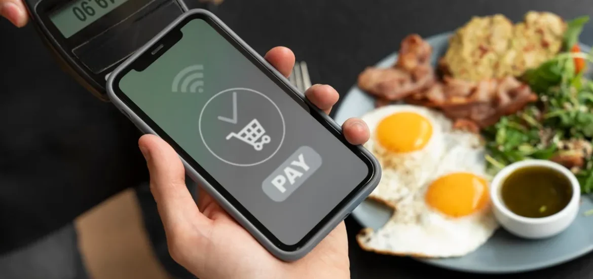 Pessoa realizando pagamento via NFC em smartphone enquanto um prato de café da manhã com ovos, bacon e salada está em segundo plano.