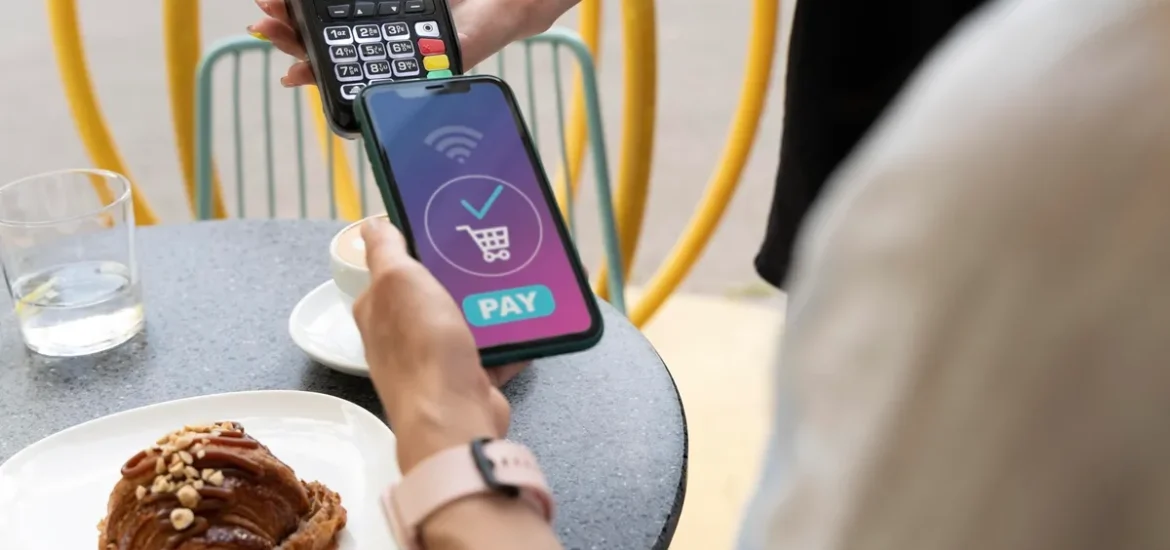 Pessoa realizando pagamento via NFC em um smartphone, próximo a uma máquina de cartão, com um croissant e café em uma mesa de café ao ar livre.