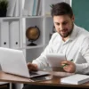 Homem analisando documentos com laptop em ambiente de trabalho organizado.