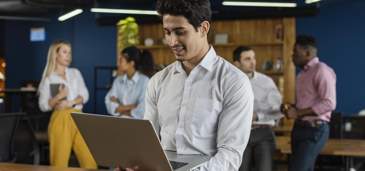 Um homem de camisa branca utiliza um notebook enquanto interage com outras pessoas em um ambiente corporativo, representando o suporte oferecido pela Sala do Empreendedor para MEIs.