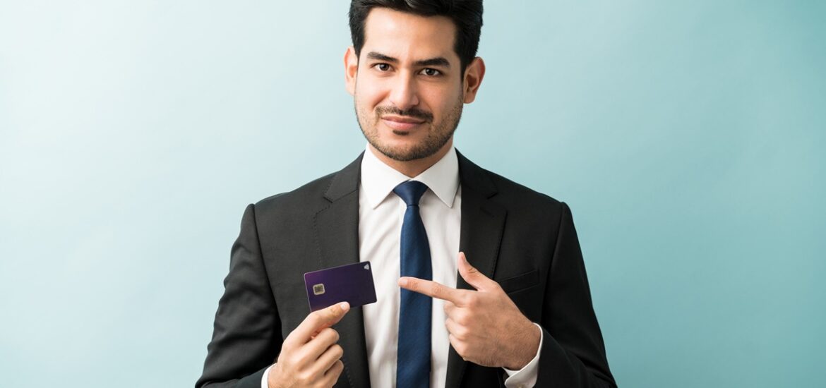 Homem de terno segurando e apontando para um cartão de crédito, com fundo azul claro.