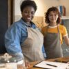 Duas mulheres empreendedoras em uma cafeteria, usando aventais, sorrindo para a câmera. Elas estão atrás de um balcão com doces expostos em recipientes de vidro, representando pequenos negócios locais.