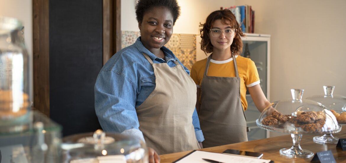 Duas mulheres empreendedoras em uma cafeteria, usando aventais, sorrindo para a câmera. Elas estão atrás de um balcão com doces expostos em recipientes de vidro, representando pequenos negócios locais.