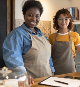 Duas mulheres empreendedoras em uma cafeteria, usando aventais, sorrindo para a câmera. Elas estão atrás de um balcão com doces expostos em recipientes de vidro, representando pequenos negócios locais.