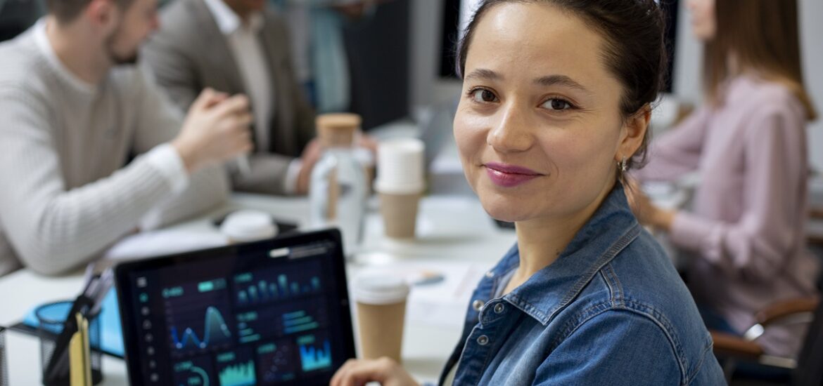 Mulher sorridente em ambiente corporativo com um laptop exibindo gráficos e dados, representando análise de negócios e empreendedorismo. Pessoas estão ao fundo em uma reunião.