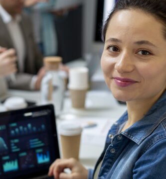 Mulher sorridente em ambiente corporativo com um laptop exibindo gráficos e dados, representando análise de negócios e empreendedorismo. Pessoas estão ao fundo em uma reunião.