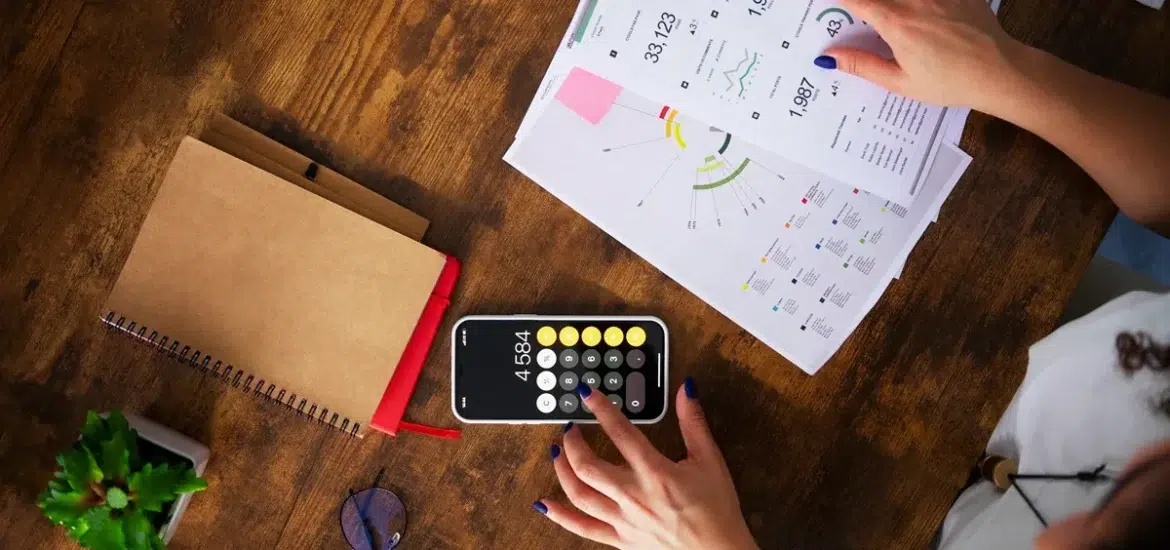 Mãos femininas analisando gráficos impressos enquanto utiliza a calculadora de um smartphone, em uma mesa de madeira com caderno, óculos e planta decorativa.