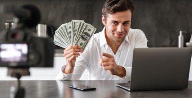 Homem segurando notas de dinheiro enquanto aponta para a câmera, com um laptop e smartphone sobre a mesa. Ele está gravando um vídeo, representando estratégias para ganhar dinheiro online.