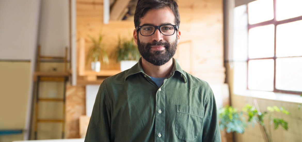 Homem sorridente com barba e óculos, vestindo camisa verde, em um ambiente iluminado com detalhes de madeira e plantas ao fundo.