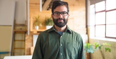 Homem sorridente com barba e óculos, vestindo camisa verde, em um ambiente iluminado com detalhes de madeira e plantas ao fundo.