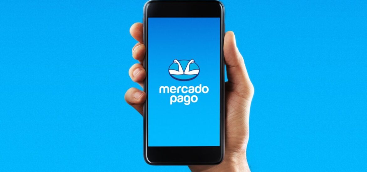 Mão segurando um smartphone com a tela aberta exibindo o logotipo do Mercado Pago sobre um fundo azul.