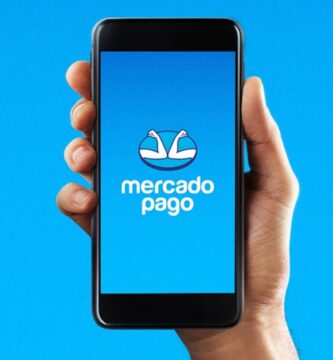 Mão segurando um smartphone com a tela aberta exibindo o logotipo do Mercado Pago sobre um fundo azul.