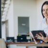 Mulher de negócios sorrindo enquanto faz split de pagamentos e analisa informações em um tablet em um ambiente de escritório moderno.