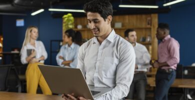 Empreendedor com laptop em mãos, trabalhando em um ambiente corporativo moderno, enquanto colegas discutem ao fundo.