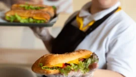 Funcionária de uma franquia da Subway segurando um sanduíche recheado com alface, tomate, queijo e pimentões, pronto para ser servido.