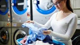 Mulher analisando roupas em uma lavanderia self-service, segurando uma peça azul enquanto retira peças de um cesto de roupas. Ao fundo, máquinas industriais de lavar e secar.
