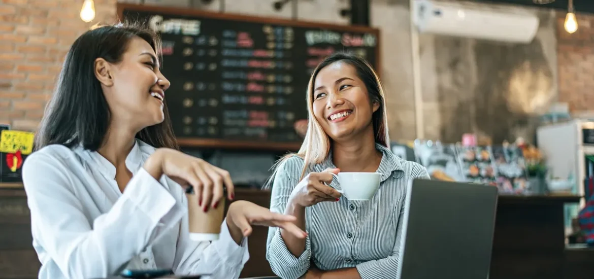 Duas mulheres sorridentes conversam em uma cafeteria aconchegante, tomando café e utilizando um laptop, representando networking e empreendedorismo em franquias.